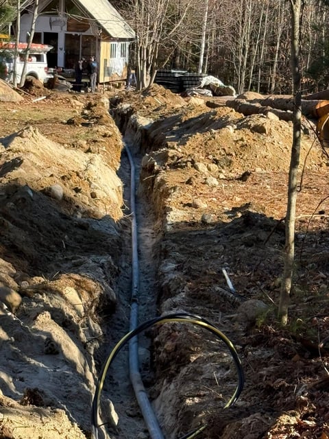 Excavated trench with blue PVC pipe and yellow cables in residential yard with wooden structure in background