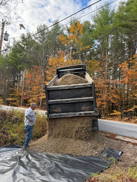 Dump truck unloading gravel and sand onto black plastic sheeting in a wooded area during autumn