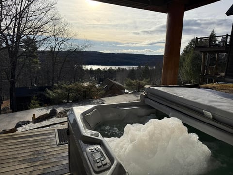 A steaming hot tub on a wooden deck overlooking a lake and forested mountains at dusk