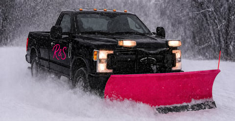 Black pickup truck with pink plow blade clearing snow during heavy snowfall