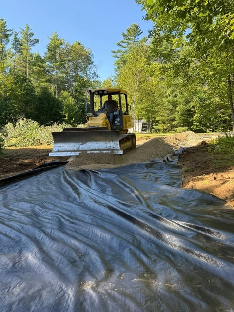 Bulldozer operating on a wooded site with geotextile fabric laid on ground, surrounded by trees and clear sky