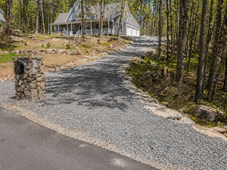 Gravel driveway leading to a cottage surrounded by pine forest, with stone pillar on left