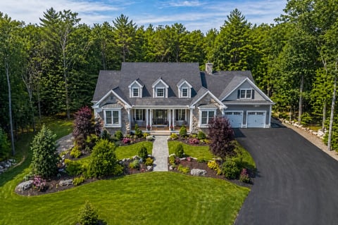 Large two-story colonial house with gray roof, front porch, and two-car garage surrounded by landscaping and trees
