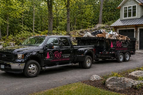 Black junk removal truck loaded with debris parked at residential driveway near wooded area